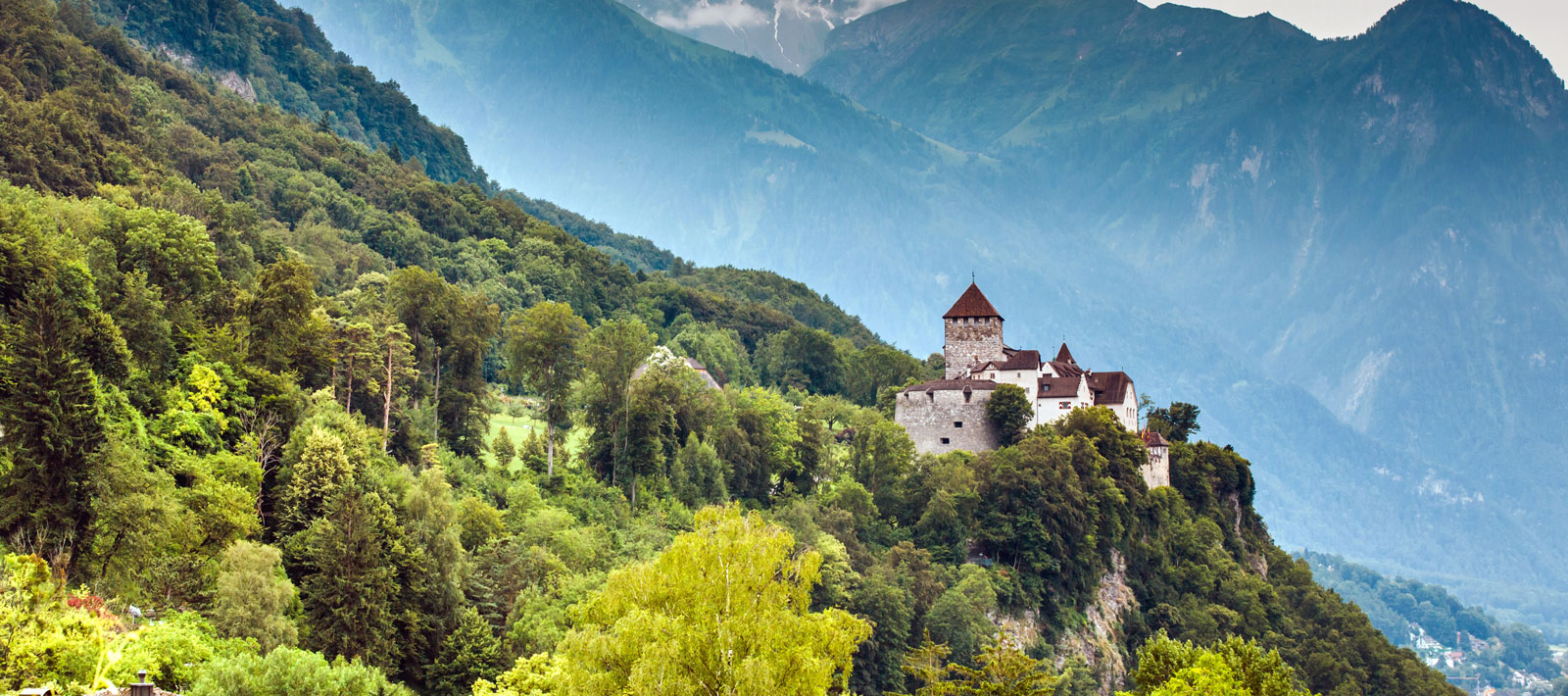 Schloss auf bewaldetem Hügel vor Bergen